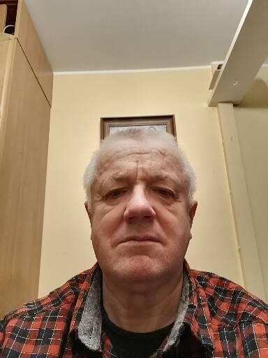 Portret mężczyzny w czerwono-czarnej koszuli w kratę na tle beżowej ściany z obrazkiem w drewnianej ramie.