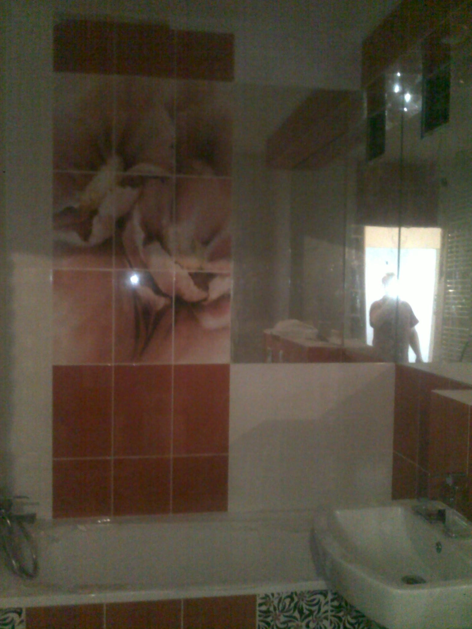 Wykończona łazienka z częściowo położonymi płytkami w odcieniach brązu i bieli, z motywem kwiatowym na ścianie i wzorzystym paskiem pod wanną, widoczne odbicie osoby w lustrze.