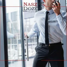 Mężczyzna w uniformie ochroniarza z napisem SECURITY na koszuli, rozmawia przez krótkofalówkę, stojąc przy przeszklonych drzwiach.