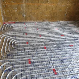 Układanie białych rur ogrzewania podłogowego na metalowej siatce z folią, widok na surowe ściany z cegieł i wystające przewody elektryczne.