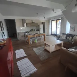 Jasne, przestronne wnętrze mieszkania z otwartą kuchnią, drewnianą podłogą i meblami w neutralnych kolorach, oświetlone wiszącymi lampami.