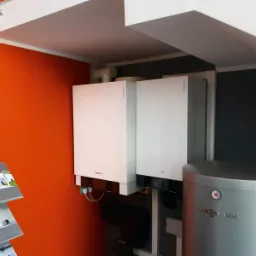 Salon firmowy Viessmann: widok na dwa białe kotły wiszące i srebrny zasobnik ciepłej wody użytkowej, na pomarańczowej ścianie ekspozytor z ulotkami.