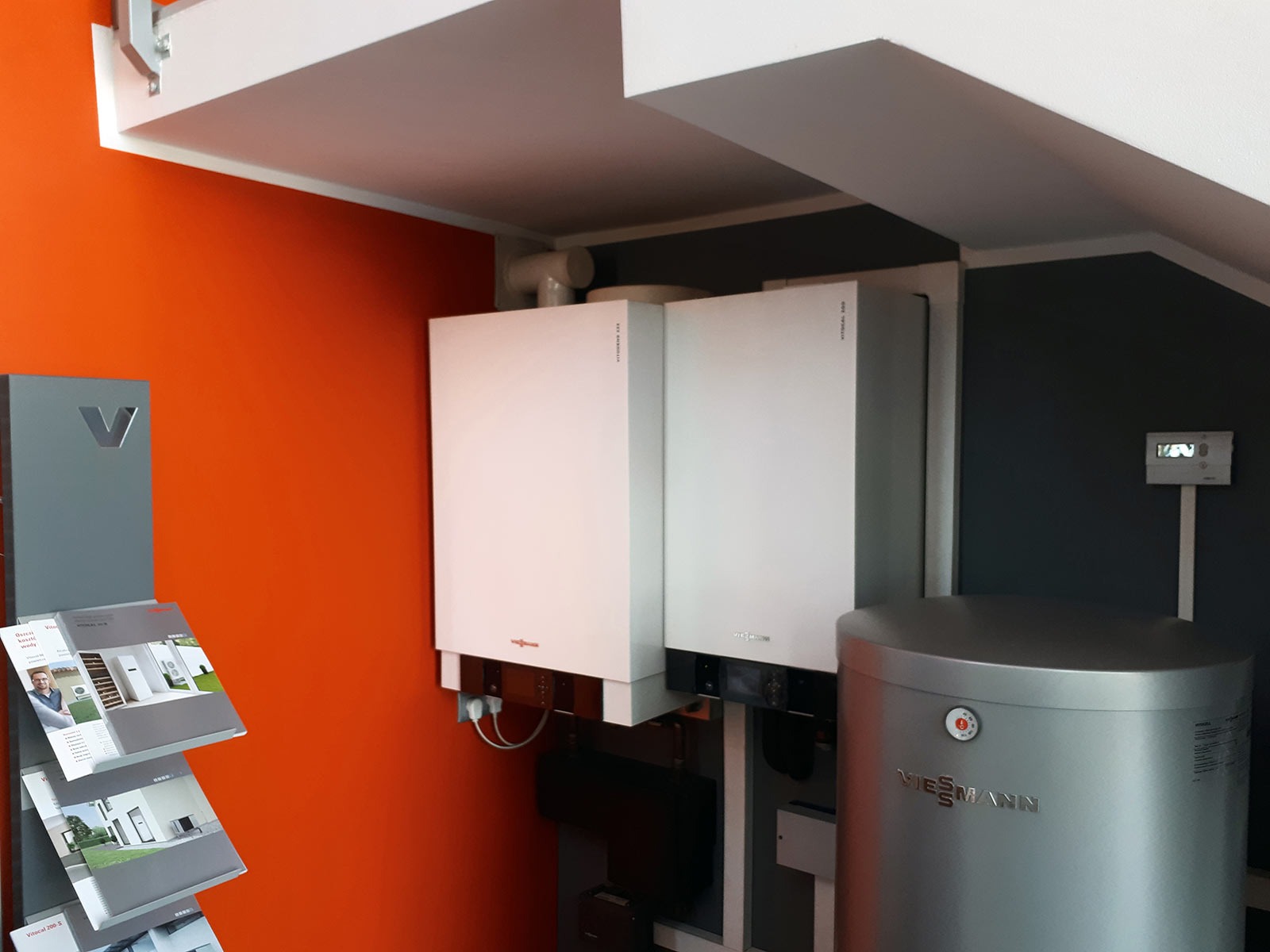 Salon firmowy Viessmann: widok na dwa białe kotły wiszące i srebrny zasobnik ciepłej wody użytkowej, na pomarańczowej ścianie ekspozytor z ulotkami.