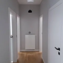 Wnętrze korytarza po odnowieniu: drewniana podłoga, szare ściany, białe drzwi z czarnymi klamkami, biały grzejnik i designerska lampa wisząca z dwoma kloszami.