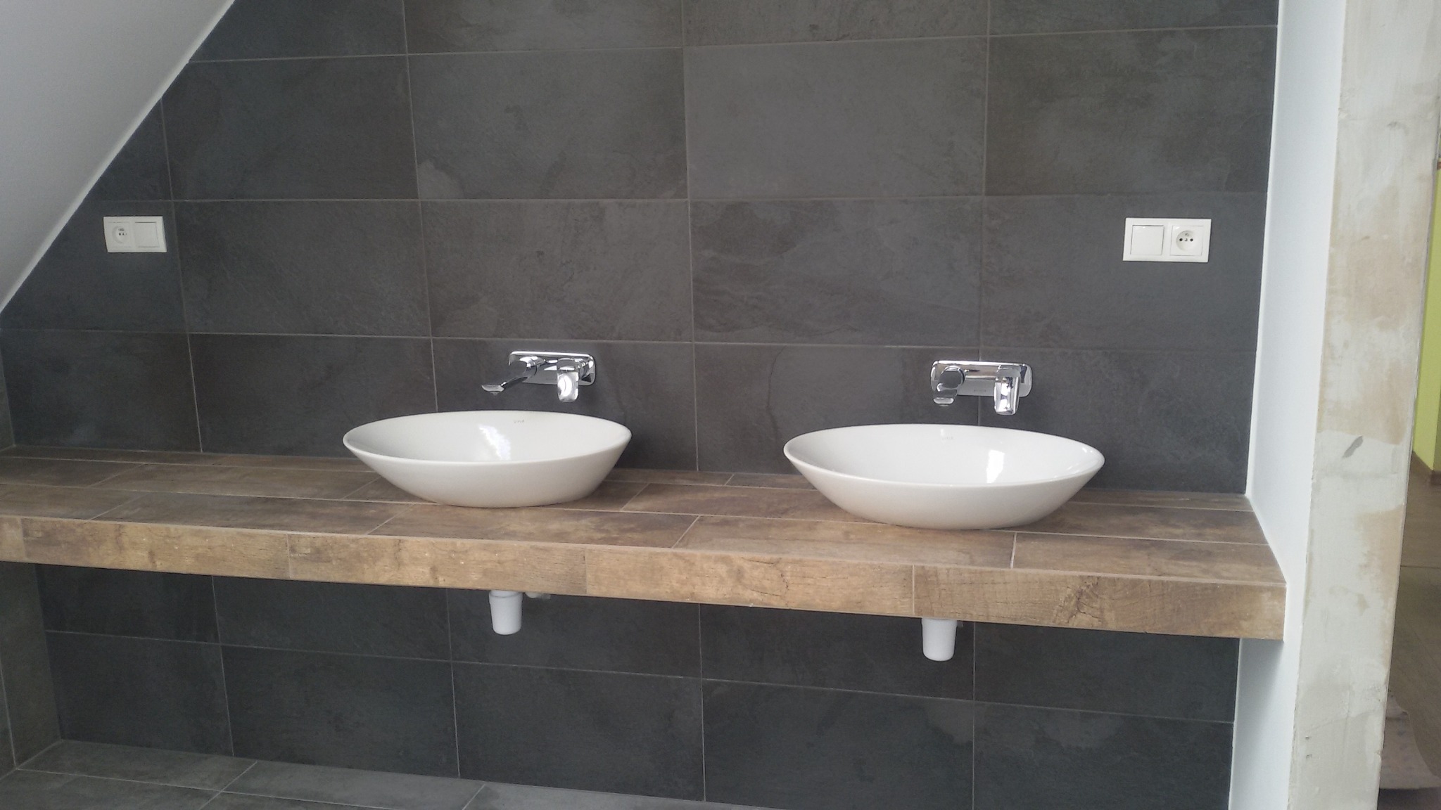 Dwie białe umywalki nablatowe z chromowanymi bateriami na blacie z płytek imitujących drewno, zamontowane na tle ciemnych, grafitowych płytek ściennych w łazience na poddaszu.