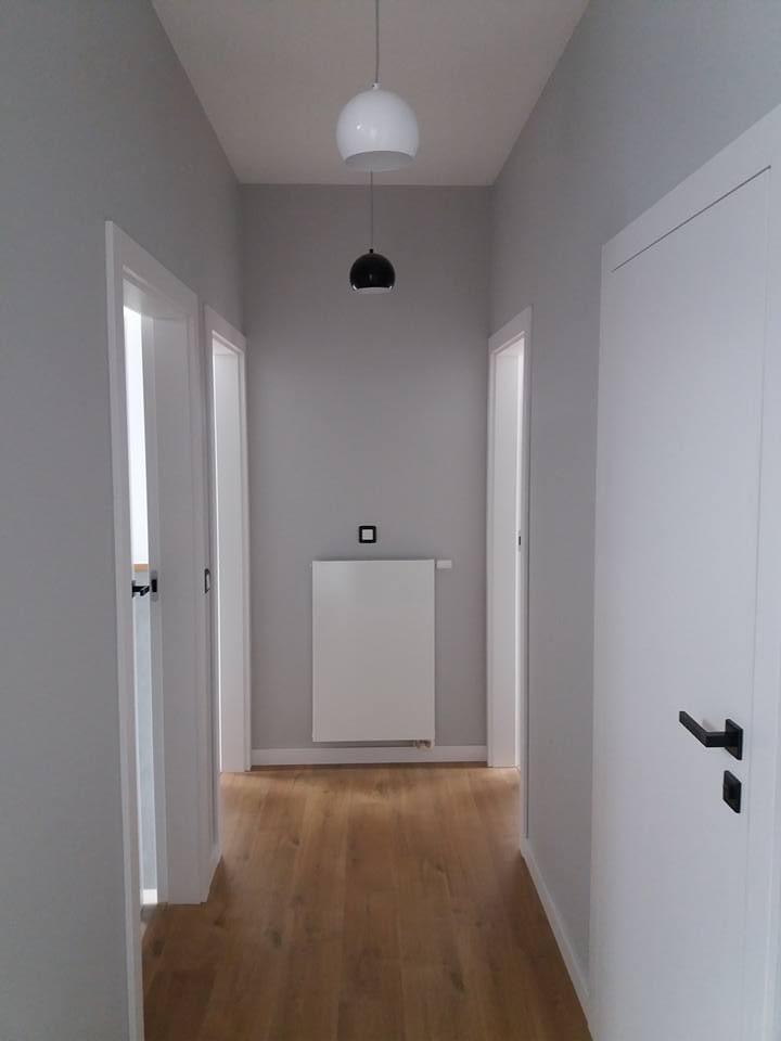 Wnętrze korytarza po odnowieniu: drewniana podłoga, szare ściany, białe drzwi z czarnymi klamkami, biały grzejnik i designerska lampa wisząca z dwoma kloszami.