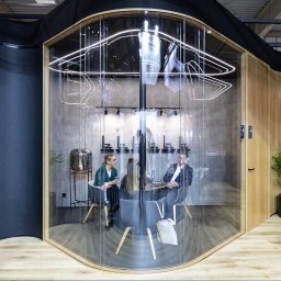 Komak - przeszklenia loftowe i biurowe - Szklana kabina biurowa z zaokrąglonymi ścianami, wewnątrz której odbywa się spotkanie. Widoczne osoby siedzące przy stole i elementy dekoracyjne.