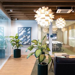 Komak - przeszklenia loftowe i biurowe - Nowoczesne biuro z przeszklonymi ścianami działowymi i roślinami doniczkowymi w zielonych donicach. Widoczne lampy wiszące i sala konferencyjna w tle.