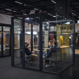 Komak - przeszklenia loftowe i biurowe - Szklana kabina biurowa z czarną ramą, wewnątrz trzech mężczyzn w trakcie rozmowy, na tle inne kabiny i stoiska targowe. Widok z boku, oświetlenie górne.
