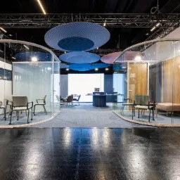 Nowoczesne, przeszklone biuro z elementami drewnianymi i okrągłymi panelami akustycznymi pod sufitem. Widoczne krzesła i stoliki w przestrzeni biurowej.