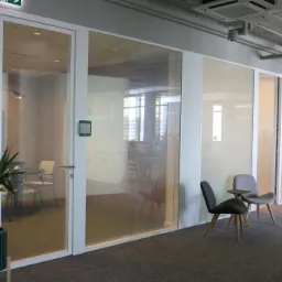 Nowoczesne biuro z przeszklonymi ścianami działowymi i drzwiami, widoczne krzesła i rośliny w donicach. Minimalistyczny design z elementami szkła.