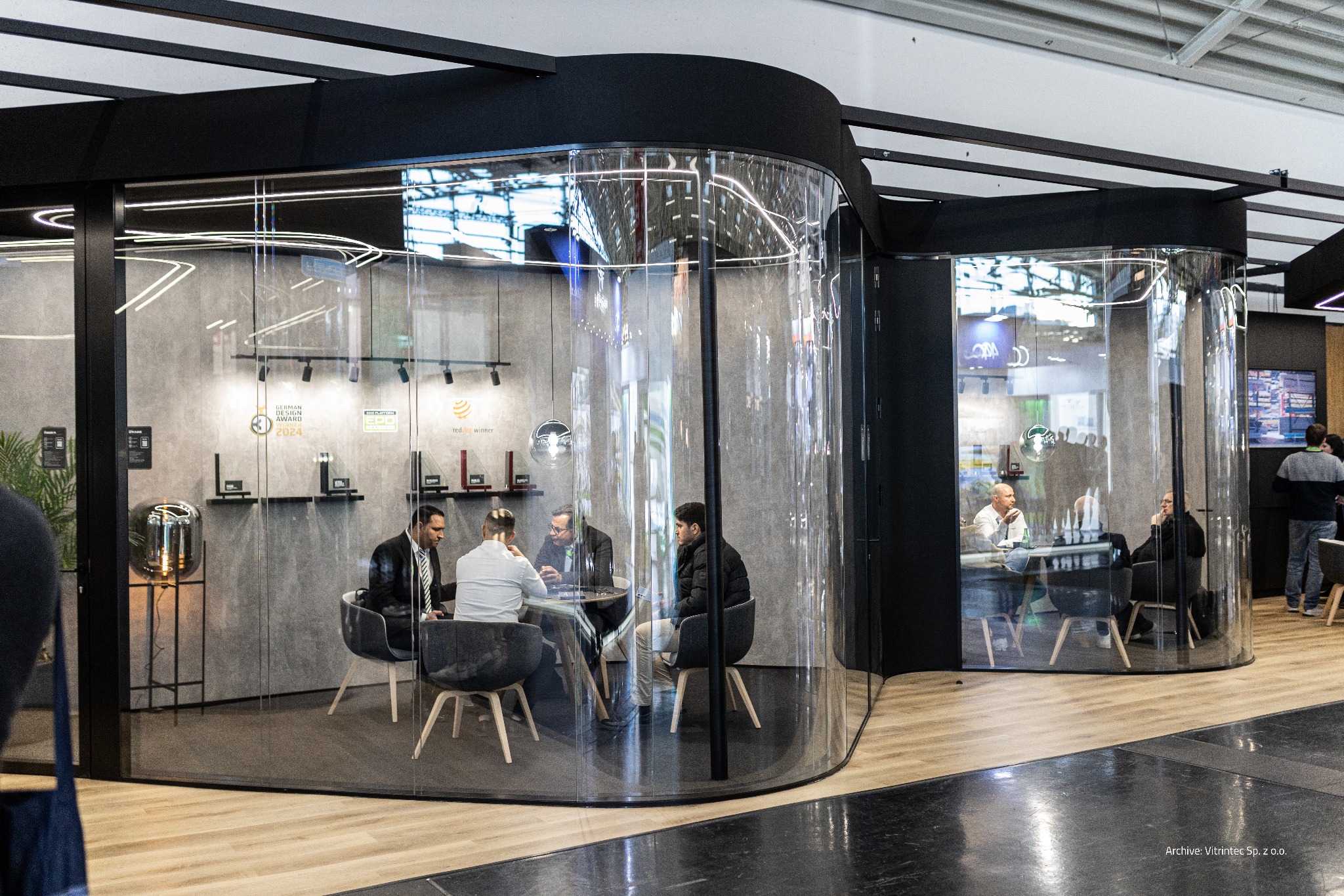 Szklana kabina konferencyjna z zaokrąglonymi ścianami, wewnątrz grupa osób przy stole. Widoczne nagrody na ścianie, oświetlenie punktowe i elementy konstrukcyjne.