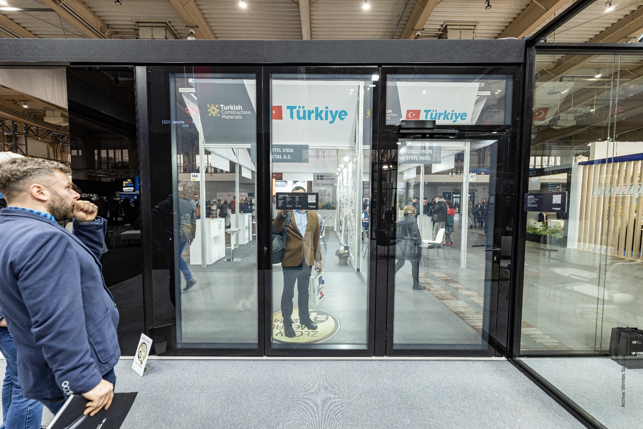 Szklane drzwi przesuwne na stoisku targowym z logo Türkiye i Turkish Construction Materials, widoczne osoby i elementy konstrukcyjne.