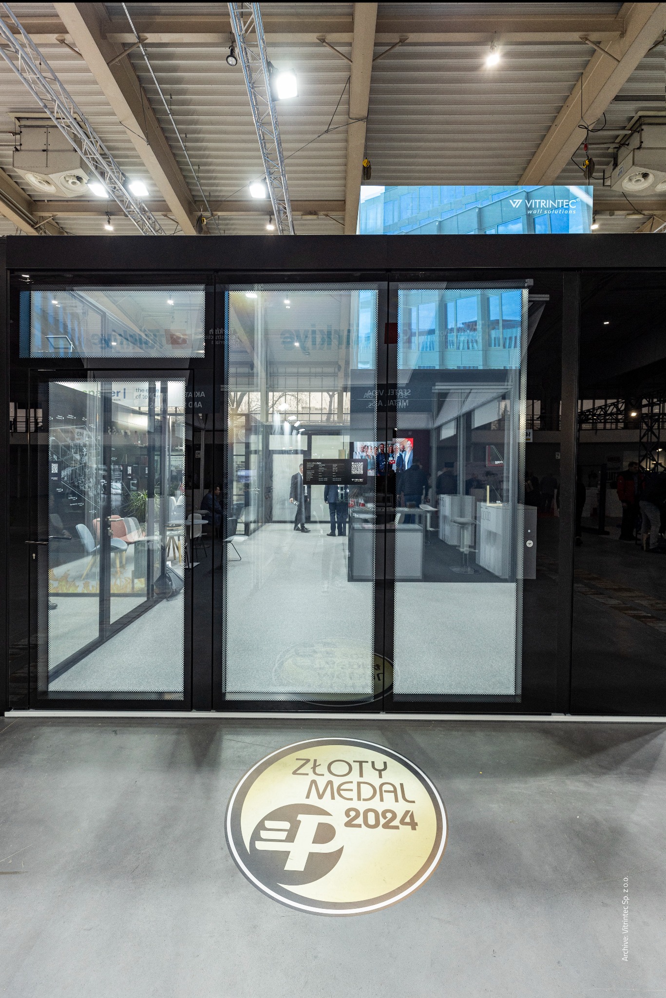 Nowoczesne drzwi przesuwne z czarnymi ramami i dużymi przeszkleniami, widoczne na tle hali targowej. Na podłodze naklejka 'Złoty Medal 2024'.