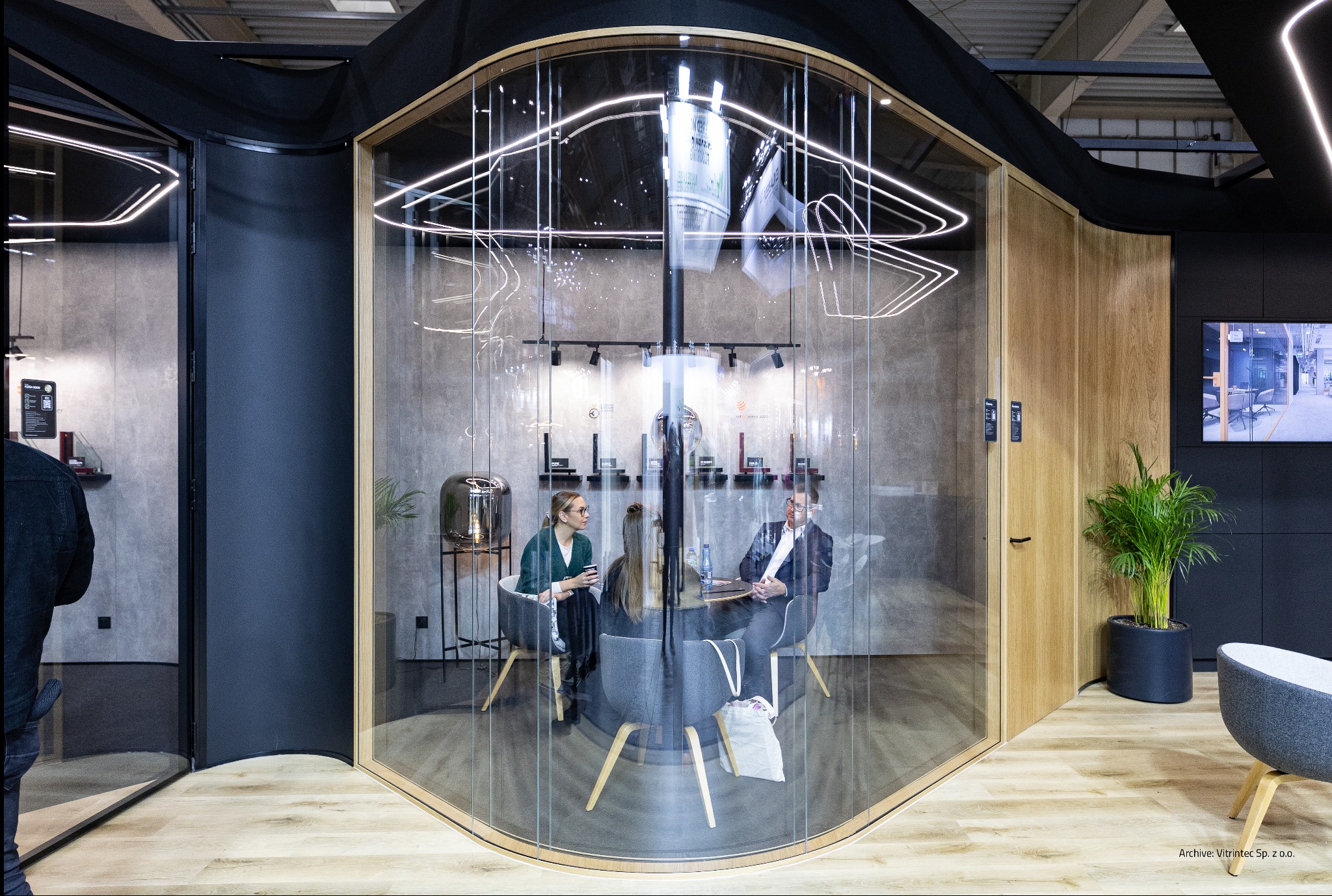 Szklana kabina biurowa z zaokrąglonymi ścianami, wewnątrz której odbywa się spotkanie. Widoczne osoby siedzące przy stole i elementy dekoracyjne.