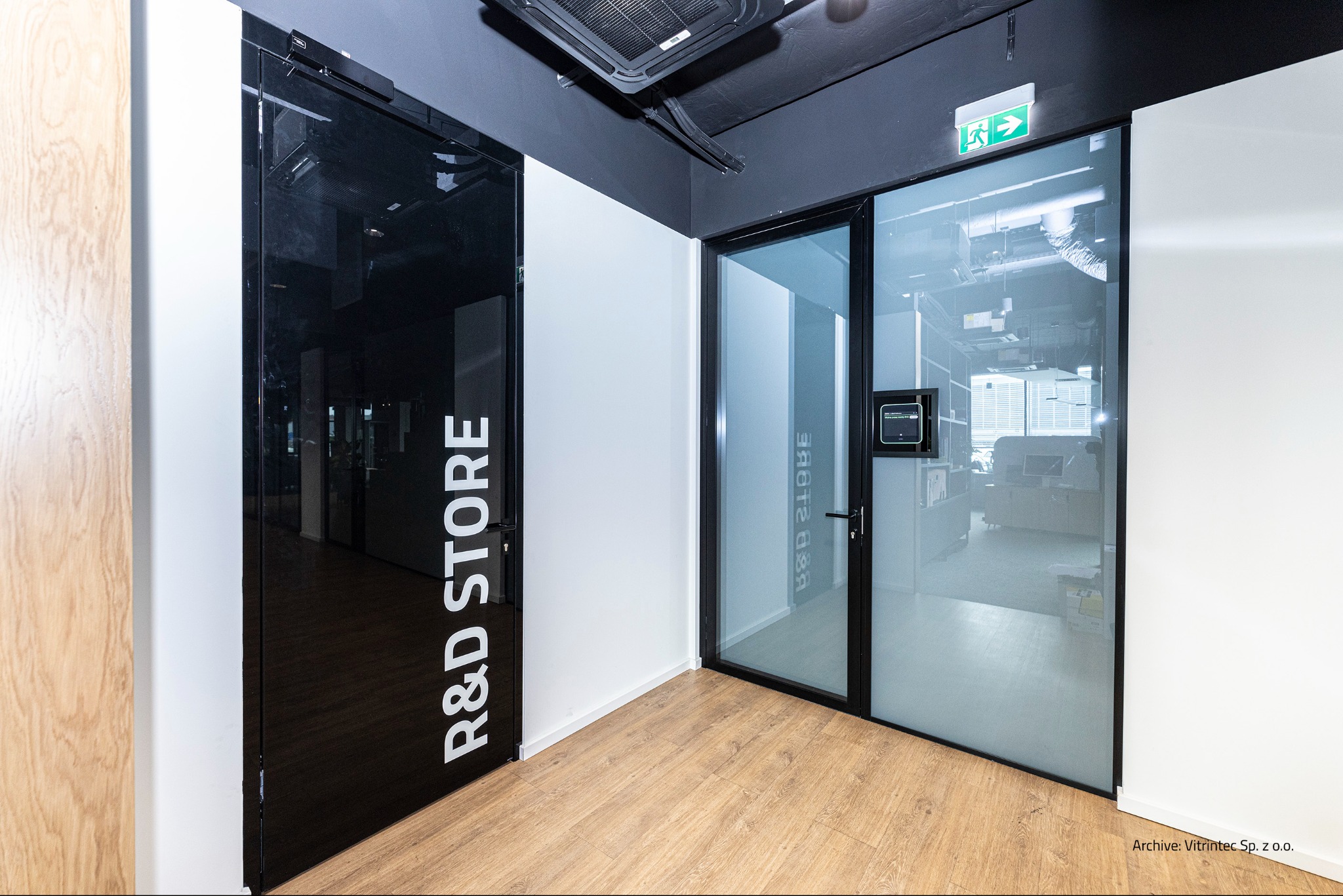 Nowoczesne, przeszklone drzwi biurowe z czarną ramą i napisem 'R&D STORE', obok drzwi szklanych z zielonym znakiem ewakuacyjnym, na jasnej podłodze.