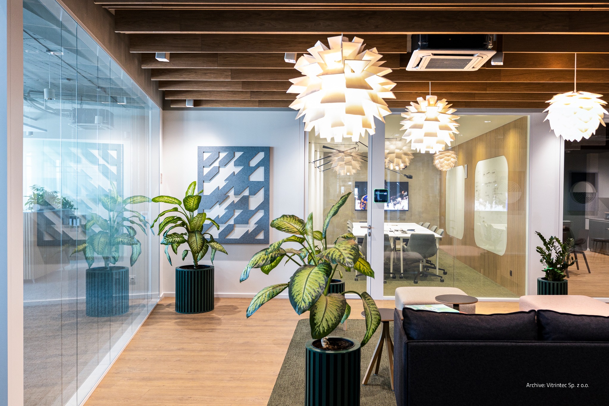 Nowoczesne biuro z przeszklonymi ścianami działowymi i roślinami doniczkowymi w zielonych donicach. Widoczne lampy wiszące i sala konferencyjna w tle.