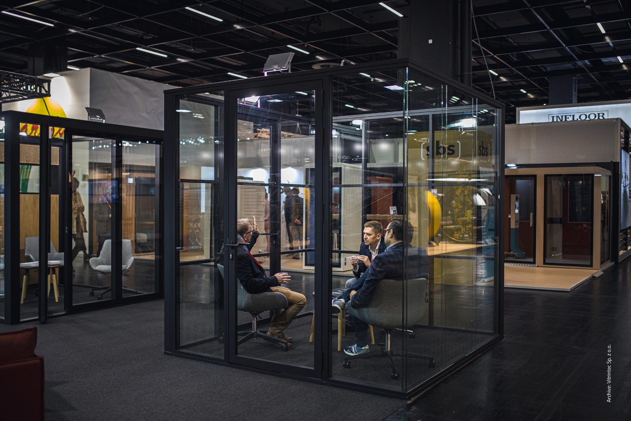 Szklana kabina biurowa z czarną ramą, wewnątrz trzech mężczyzn w trakcie rozmowy, na tle inne kabiny i stoiska targowe. Widok z boku, oświetlenie górne.