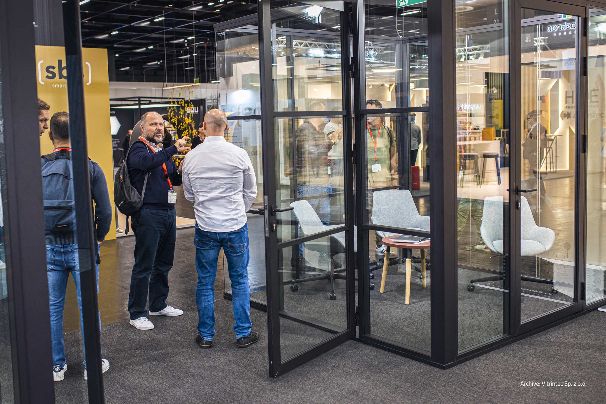 Szklana kabina biurowa z czarnymi ramami, otwarte drzwi, widoczni ludzie w tle, krzesła biurowe w środku, oświetlenie sufitowe, ekspozycja targowa.