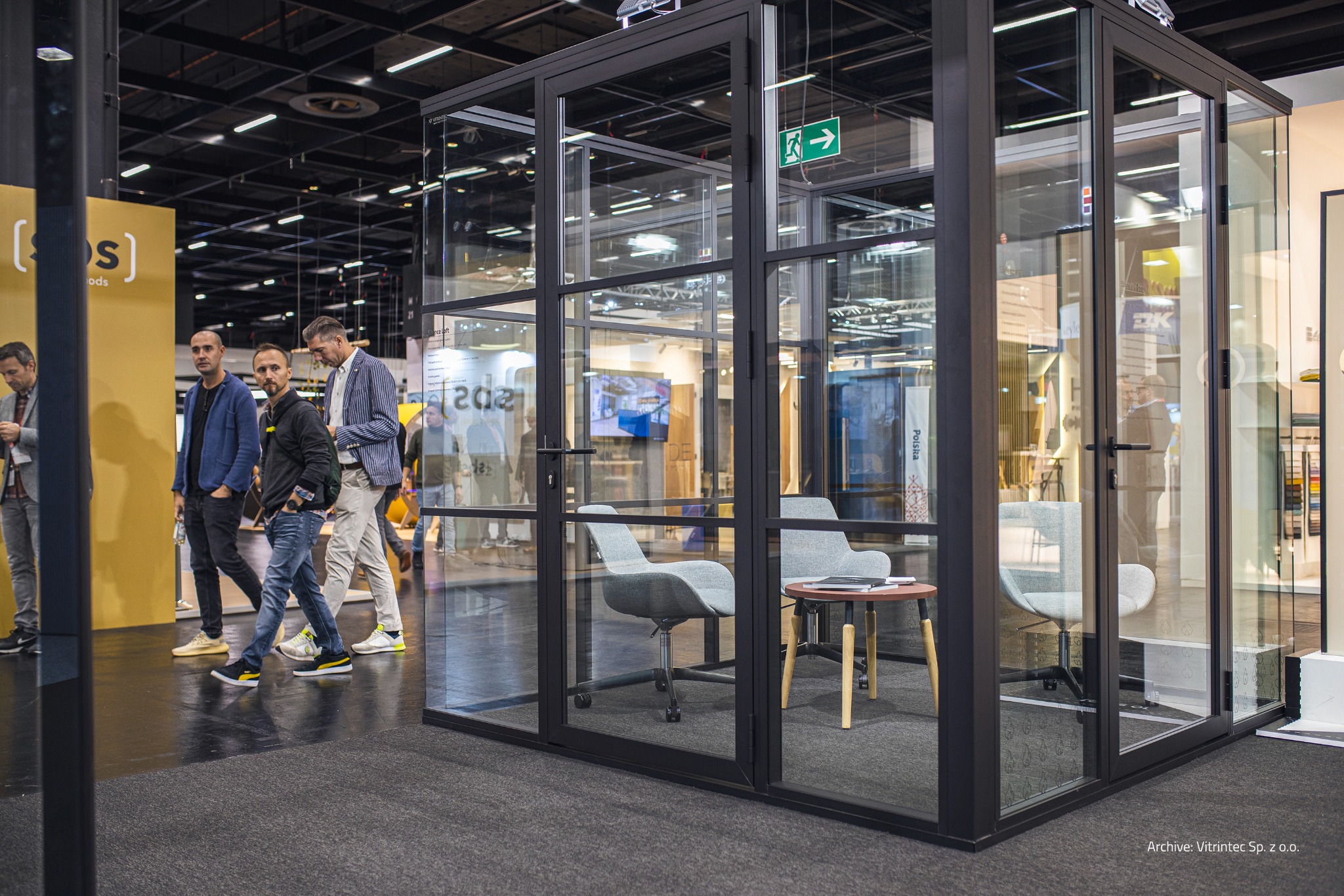 Szklana kabina biurowa z czarną ramą, widoczna na tle targowym z przechodzącymi osobami. Wnętrze wyposażone w krzesła i stolik. Oświetlenie sufitowe.