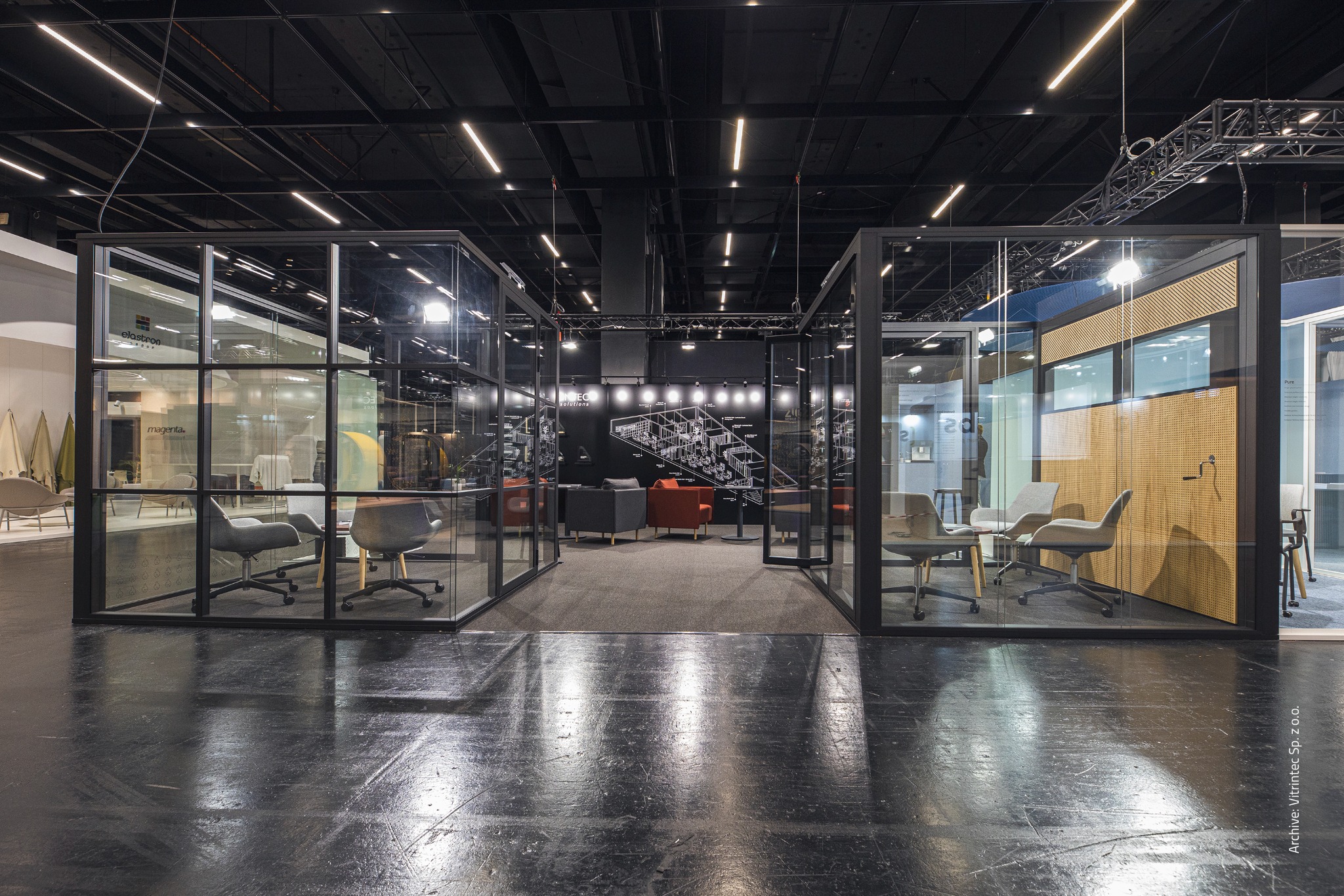 Szklane ścianki działowe z czarnymi ramami tworzą otwarte przestrzenie biurowe z widocznymi krzesłami i panelami akustycznymi. Nowoczesny design, oświetlenie sufitowe.