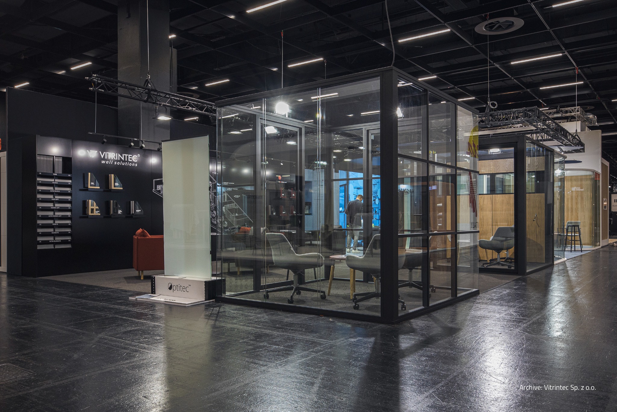 Szklana zabudowa biurowa z czarnymi ramami na targach, oświetlenie sufitowe, widoczne krzesła biurowe wewnątrz. Marka Vitrintec w tle.