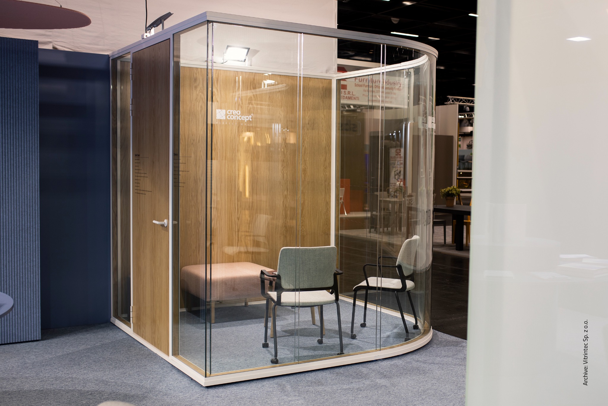 Szklana kabina biurowa z drewnianym wnętrzem, wyposażona w siedziska i krzesła. Kabina ma zaokrągloną ścianę ze szkła giętego.