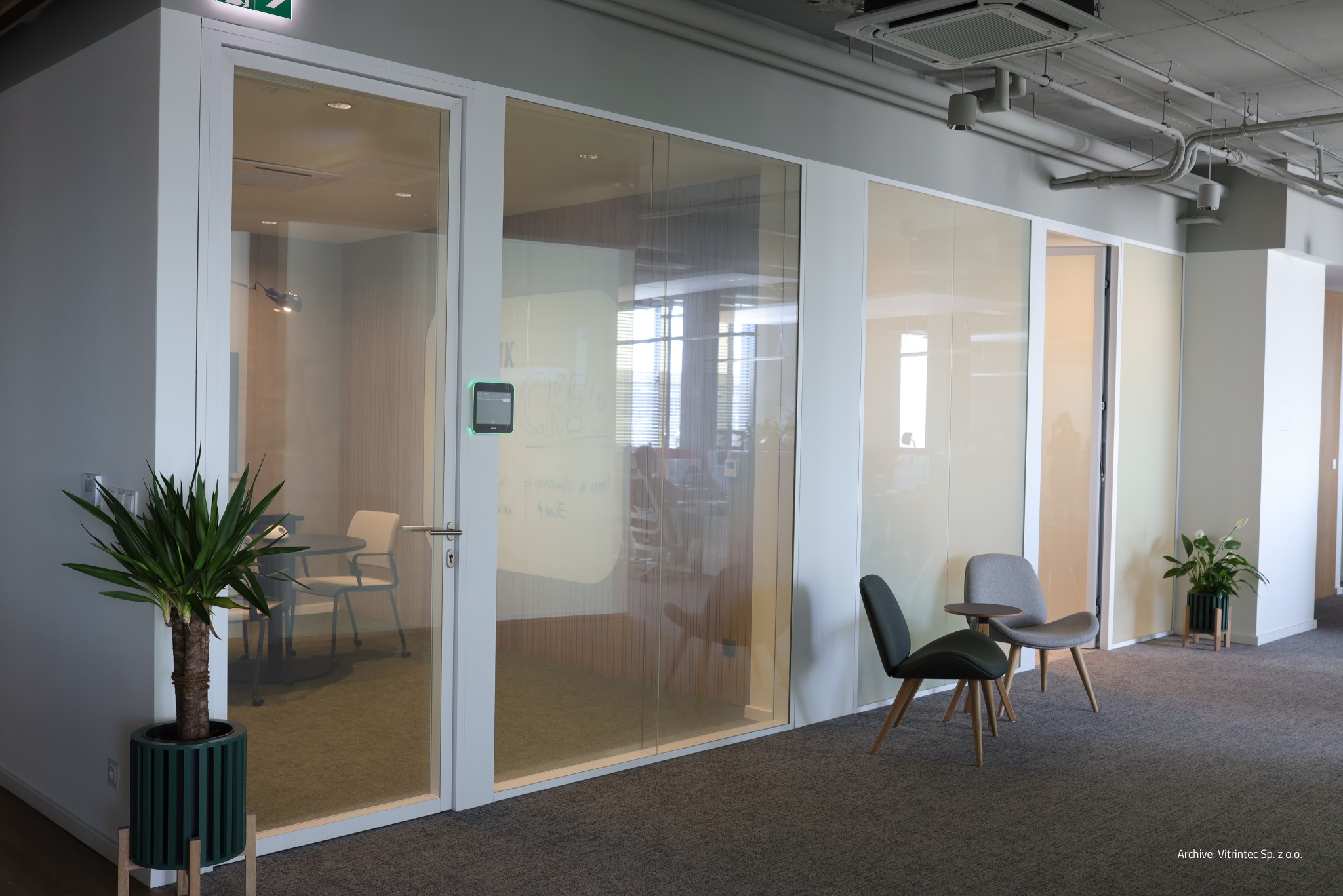 Nowoczesne biuro z przeszklonymi ścianami działowymi i drzwiami, widoczne krzesła i rośliny w donicach. Minimalistyczny design z elementami szkła.