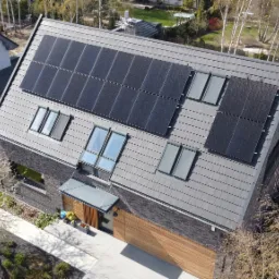 Widok z góry na nowoczesny dom z szarej cegły z panelami słonecznymi na dachu, obok kwitnące drzewo i inny dom.