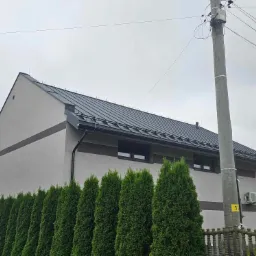 Nowoczesny dom z szarą blachodachówką, widoczne rynny i rząd tuj, słup elektryczny z numerem 32 na pierwszym planie.