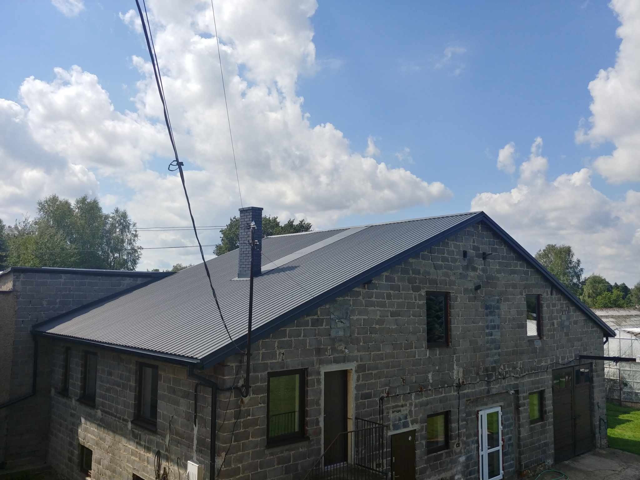 Szary budynek z nowym, metalowym dachem, widoczne komin z cegły klinkierowej, rynny w kolorze dachu i fragment instalacji odgromowej na tle błękitnego nieba z chmurami.