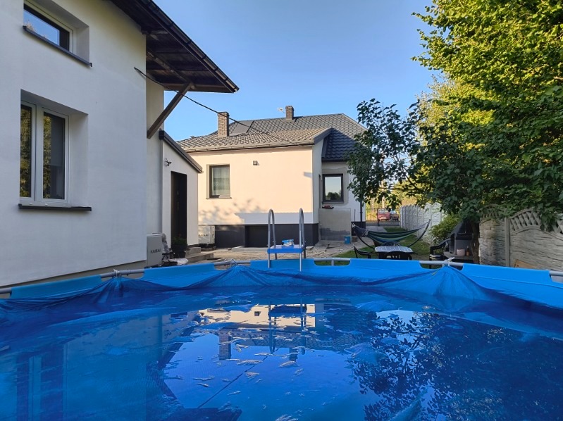 Nowoczesny dom z ciemnoszarym dachem z dachówki ceramicznej, widoczny z perspektywy basenu ogrodowego z niebieską plandeką.