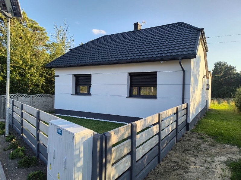 Nowoczesny dom z ciemną, metalową dachówką, widoczne okna z roletami zewnętrznymi i betonowe ogrodzenie panelowe.