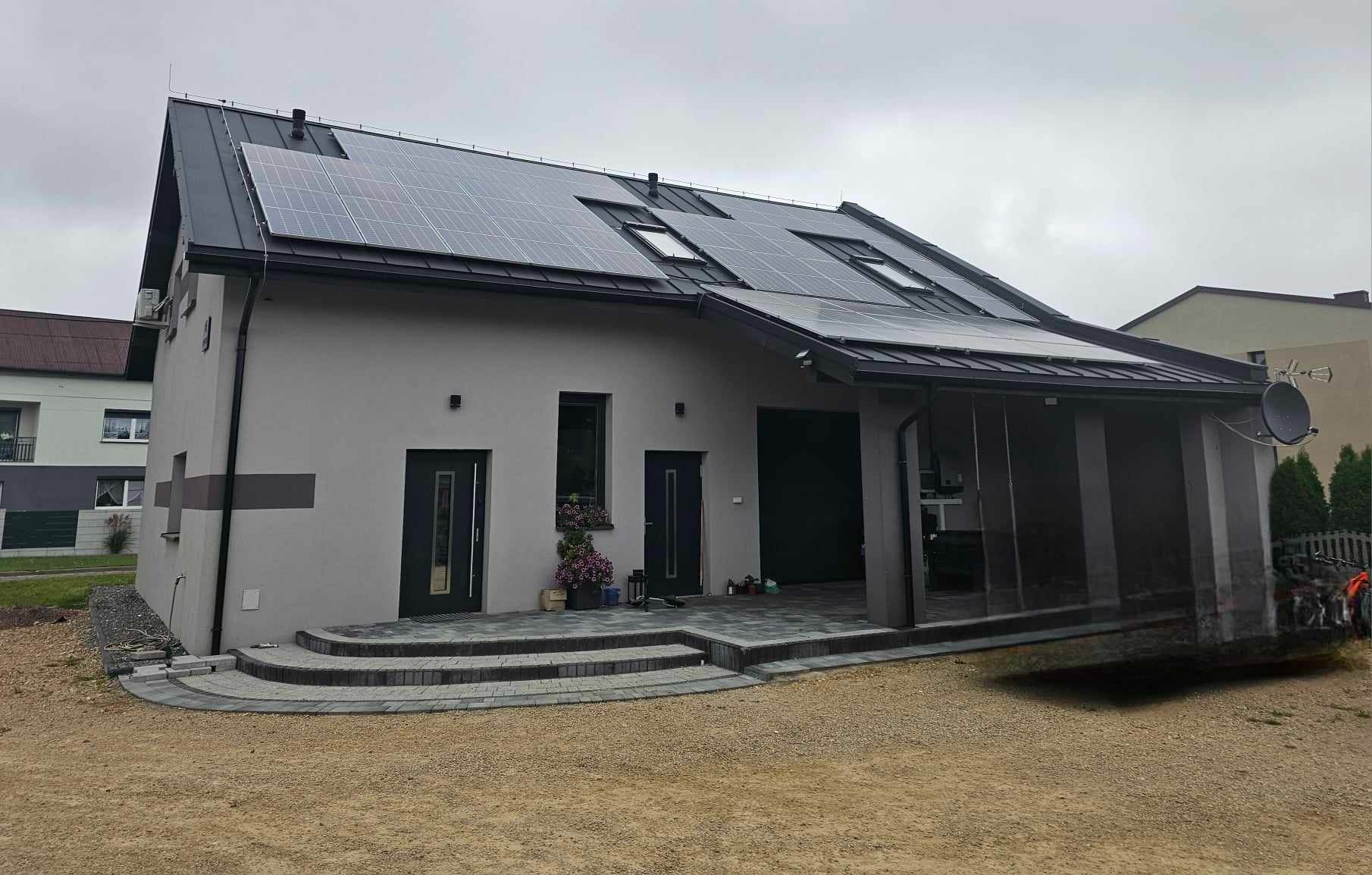 Szary dom jednorodzinny z ciemnym dachem, panelami słonecznymi i oknami dachowymi, widok z przodu na elewację i wejście z kostki brukowej.