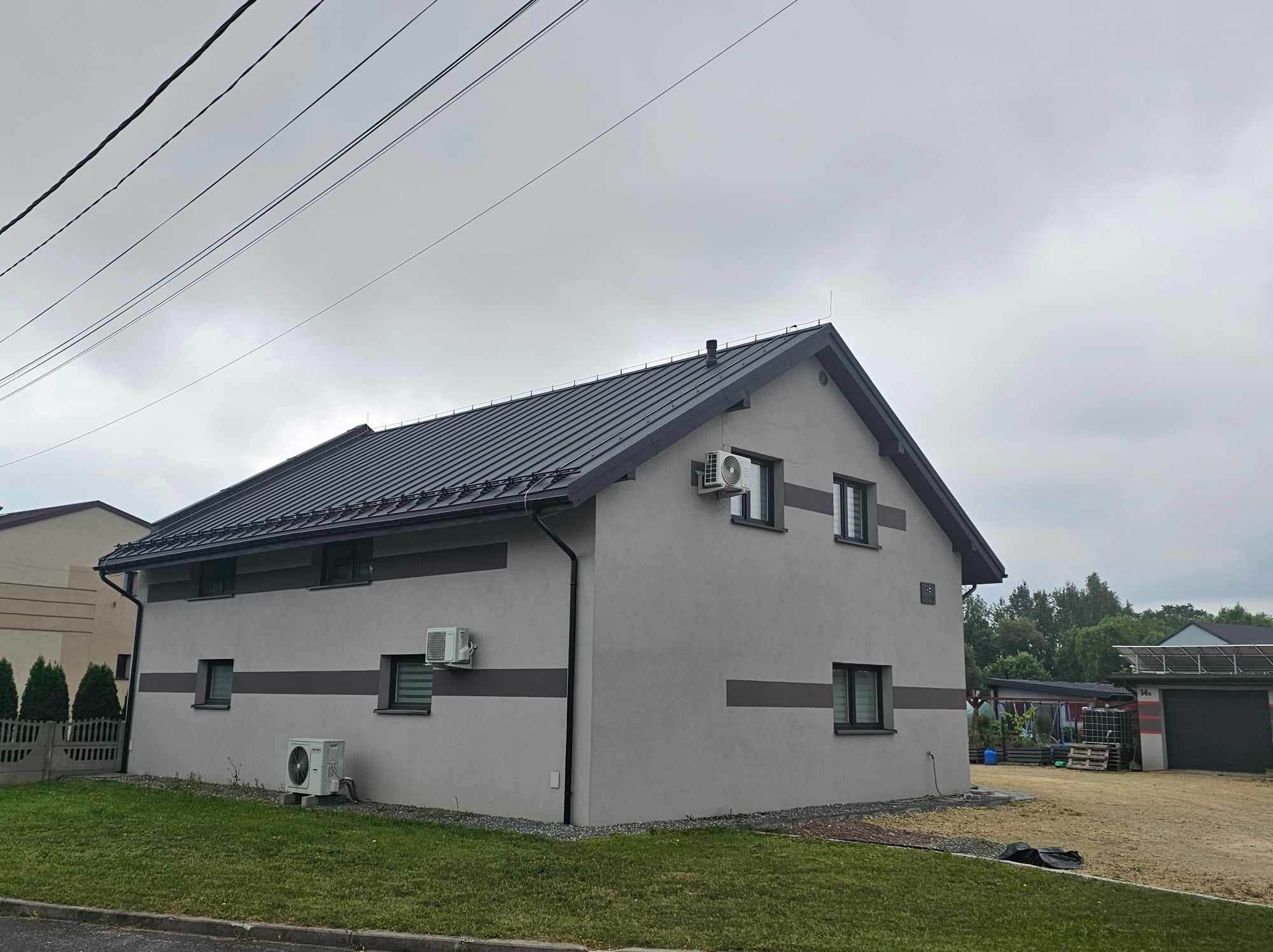 Dwukondygnacyjny dom z szarą, metalową blachą na dachu, widoczne rynny w kolorze dachu, trzy klimatyzatory na elewacji, szara elewacja z ciemniejszymi pasami, trawnik przed budynkiem, pochmurne niebo.