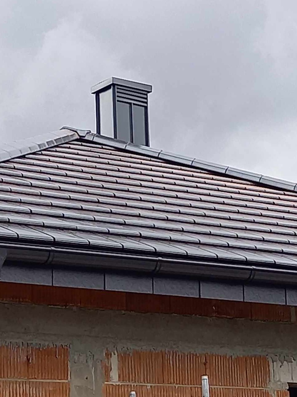 Szary dach z dachówki ceramicznej z nowoczesnym kominem wentylacyjnym na tle pochmurnego nieba, widoczne elementy muru w stanie surowym.