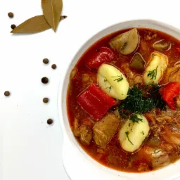 Porcja gęstej zupy gulaszowej w białej misce, z ziemniakami, papryką i pieczarkami, udekorowana koperkiem, z liśćmi laurowymi i ziarnami pieprzu na białym tle.