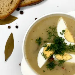 Żurek w białym talerzu z połówkami jajka i koperkiem, obok kromki ciemnego chleba, liść laurowy i ziarna pieprzu na białym tle.