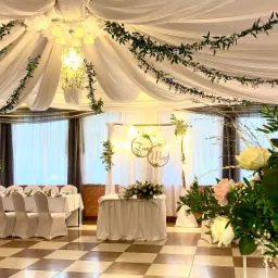 Uroczysta sala bankietowa z dekoracją z białych draperii, girland z zielonych liści, kryształowego żyrandola i kompozycji kwiatowych z białych i różowych róż. W tle ustawione stoły z białymi...