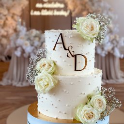 FUNDACJA AMBROZJA - Dwupiętrowy tort weselny w kremowej tonacji, zdobiony białymi różami i perłowymi koralikami, z inicjałami 'A&D' na tle dekoracji kwiatowych i napisu.