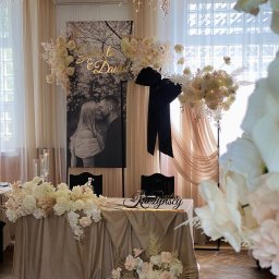 FUNDACJA AMBROZJA - Elegancko udekorowany stół weselny z kwiatami w odcieniach bieli i beżu, za nim czarno-białe zdjęcie pary młodej i złoty napis. Całość w jasnej, przestronnej sali.