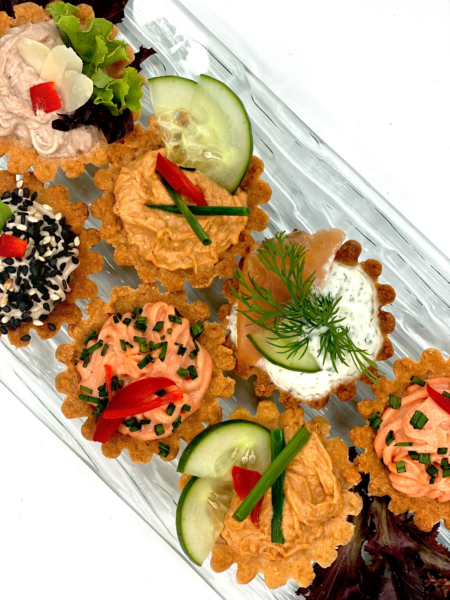 Szklany talerz z siedmioma tartaletkami na kruchym spodzie, każda z innym nadzieniem: pasta rybna z ogórkiem i koperkiem, pasta z łososiem, pasta z sezamem, pasta z warzywami i ziołami, udekorowane...