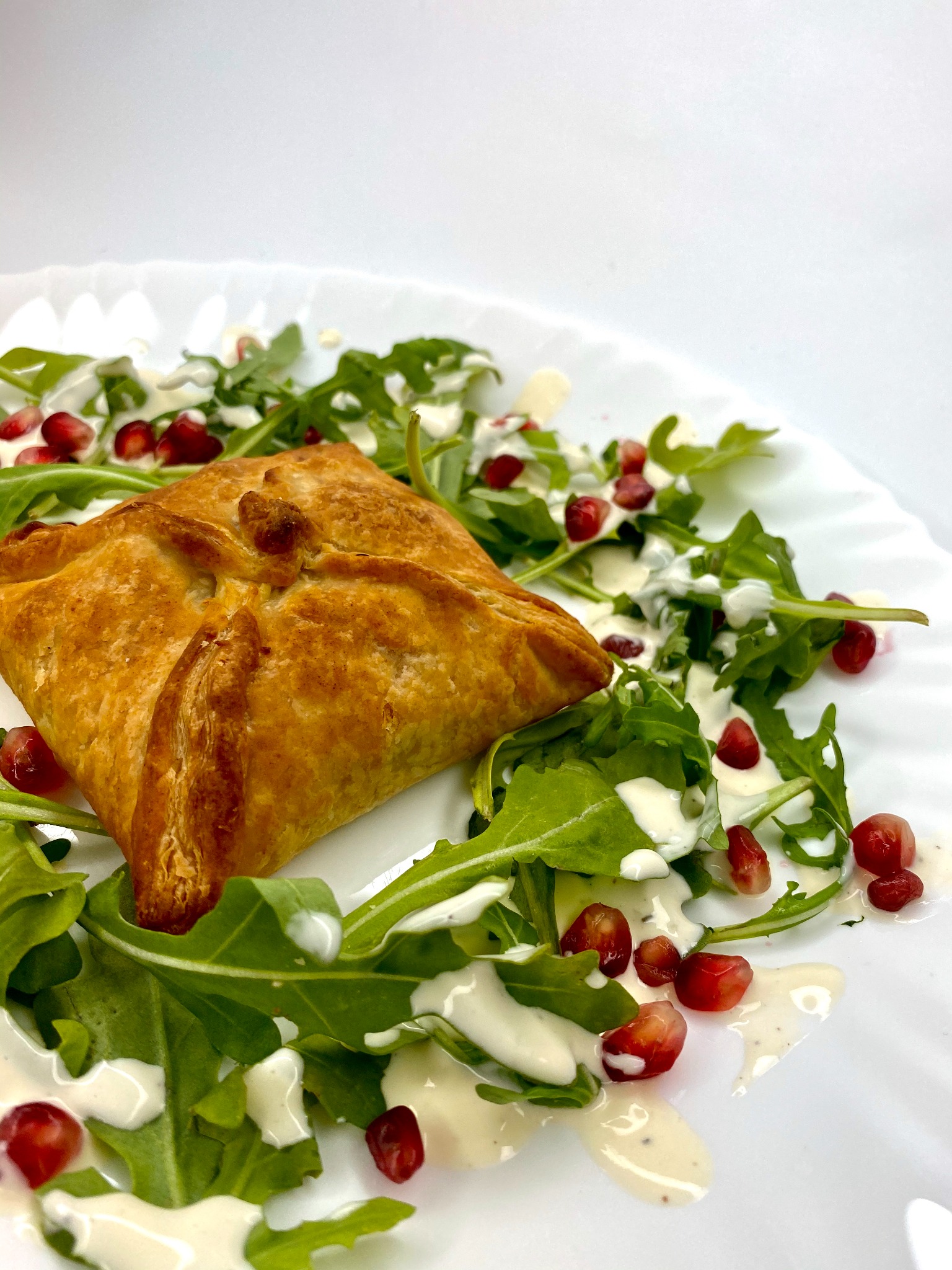 Złocisty pieróg z ciasta francuskiego, ułożony na białym talerzu z rukolą, sosem i pestkami granatu.