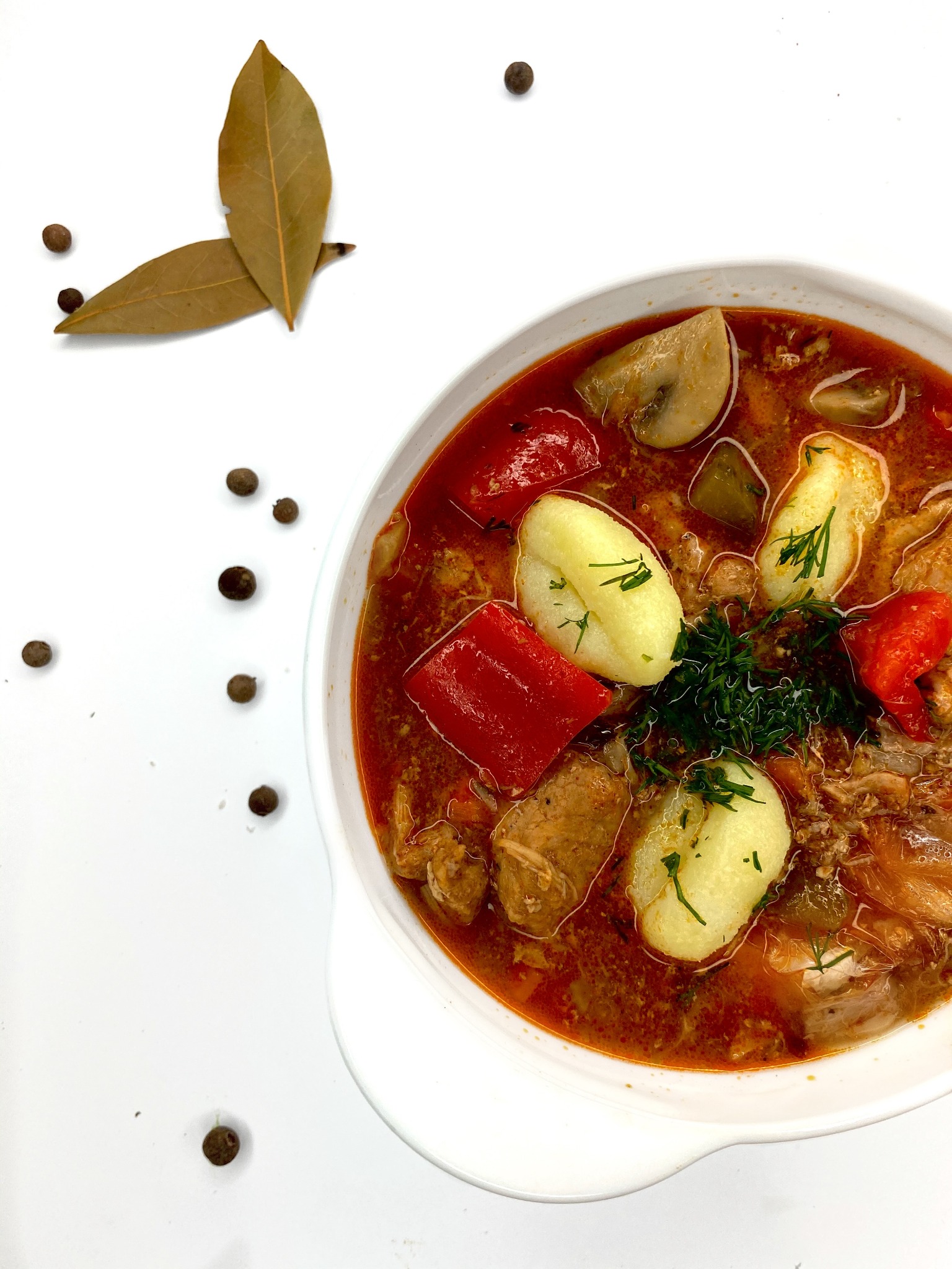 Porcja gęstej zupy gulaszowej w białej misce, z ziemniakami, papryką i pieczarkami, udekorowana koperkiem, z liśćmi laurowymi i ziarnami pieprzu na białym tle.