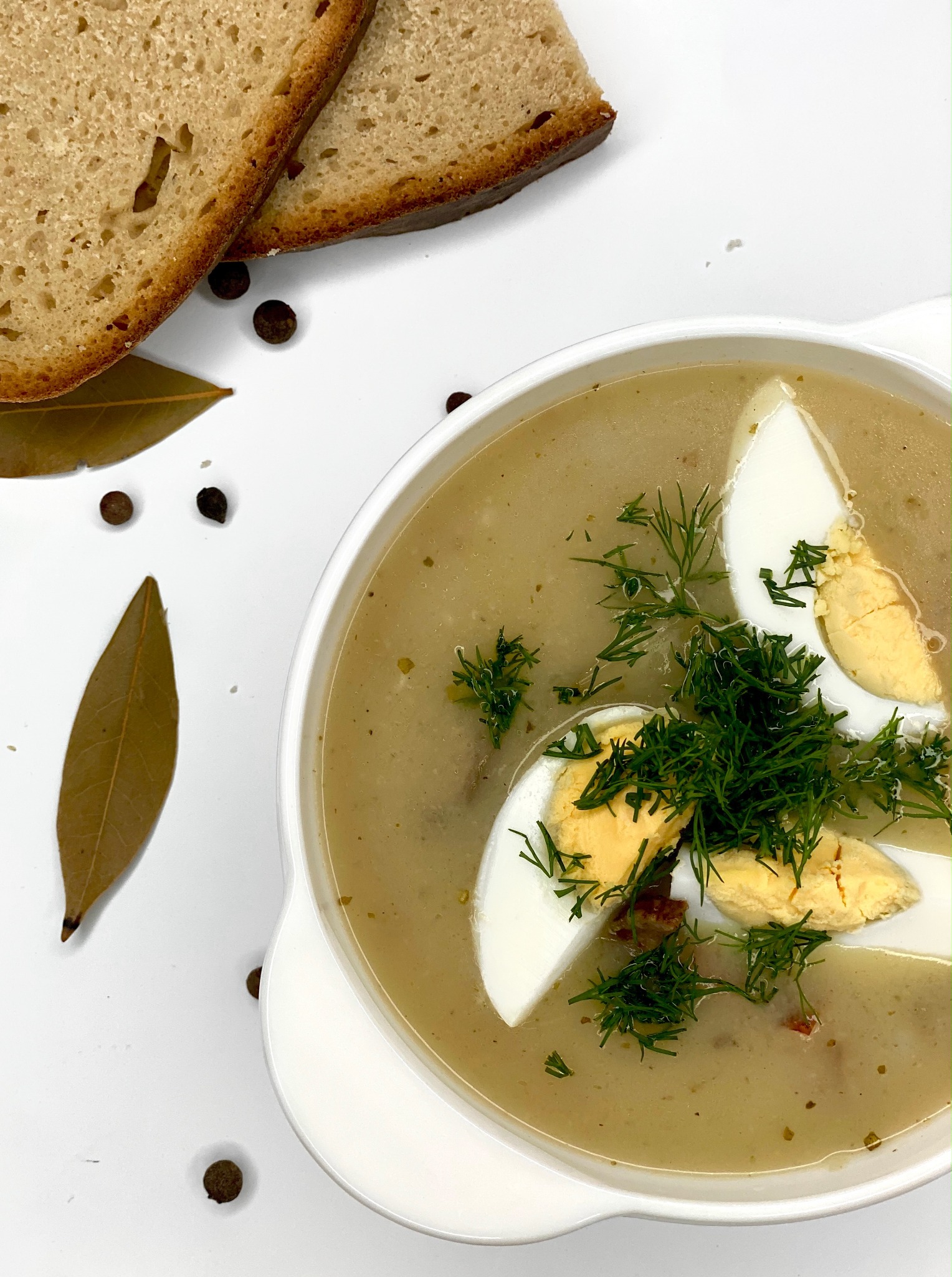 Żurek w białym talerzu z połówkami jajka i koperkiem, obok kromki ciemnego chleba, liść laurowy i ziarna pieprzu na białym tle.