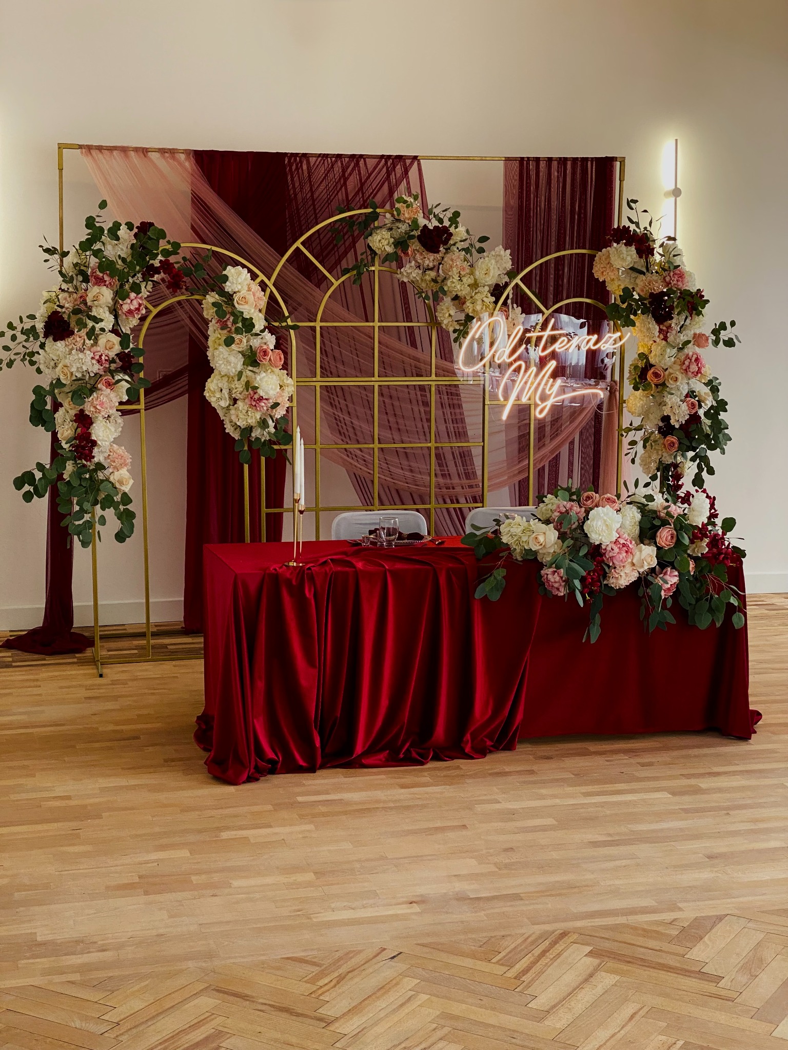 Uroczysty stół weselny z czerwonym obrusem, kwiatowymi dekoracjami i neonowym napisem 'Od teraz My' na złotym stelażu. Elegancka aranżacja sali.