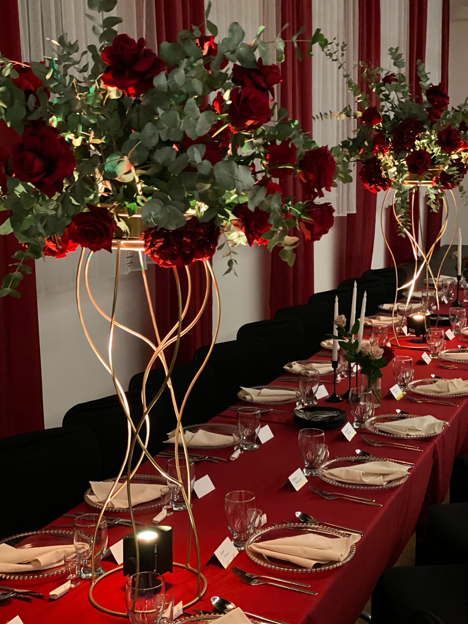 Uroczysty stół z czerwonym obrusem, elegancką zastawą i wysokimi kompozycjami z czerwonych róż w złotych wazonach. Dekoracja eventowa z dbałością o detale.