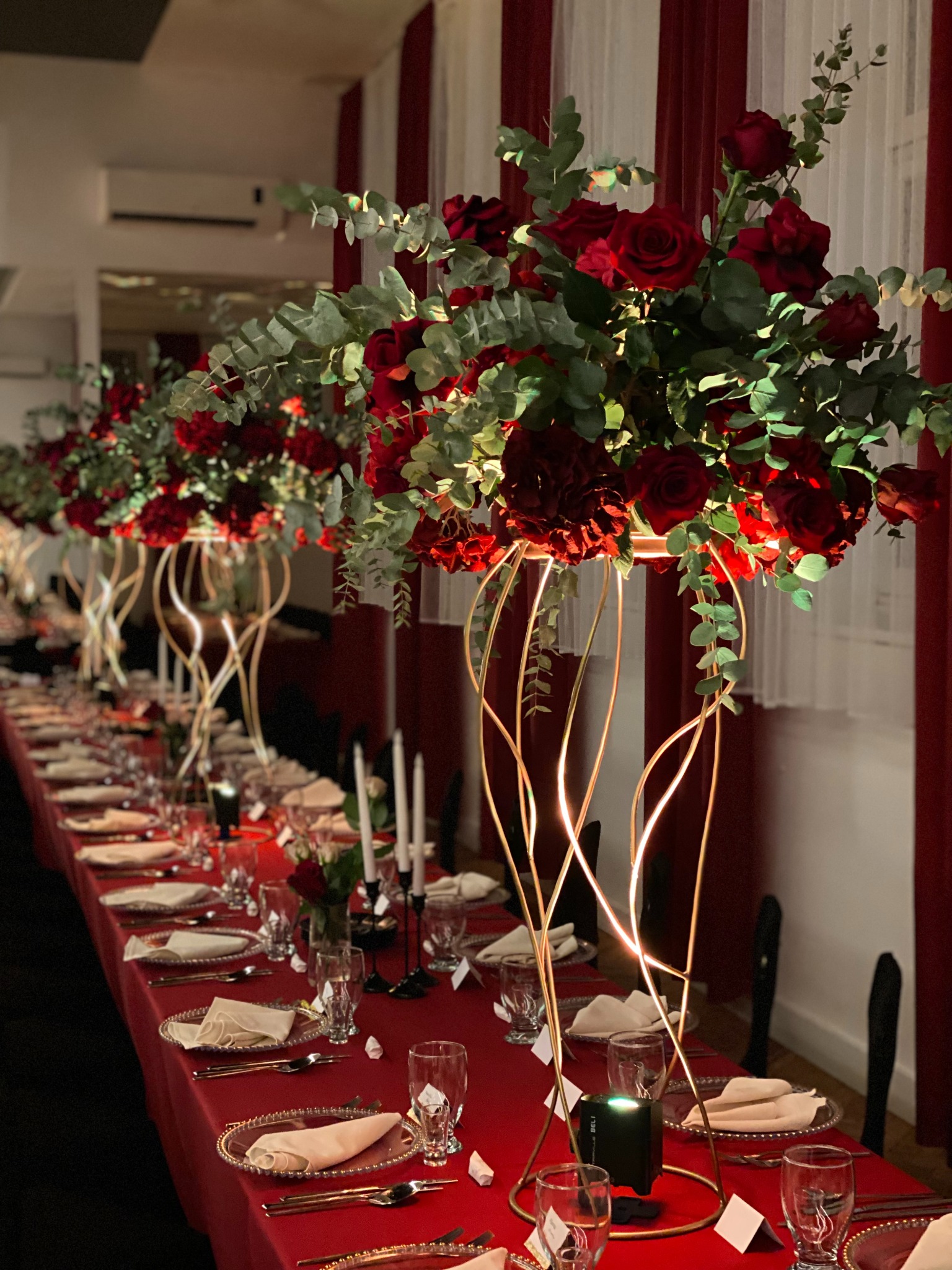 Uroczysty stół z czerwonym obrusem, nakryciami i wysokimi kompozycjami z czerwonych róż i eukaliptusa w złotych, podświetlanych wazonach. Elegancka aranżacja.