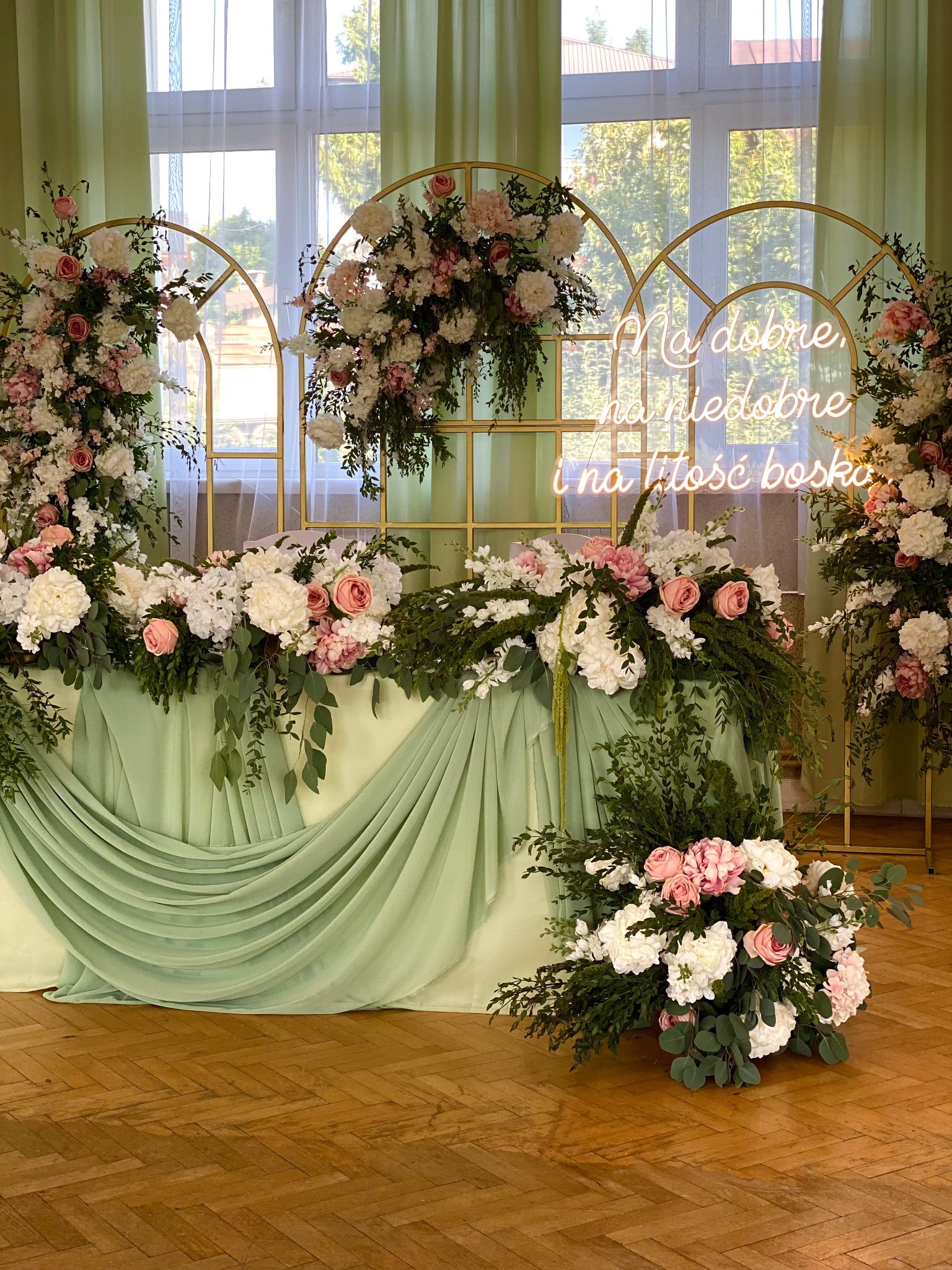 Dekoracja stołu weselnego z kwiatami w odcieniach bieli i różu, podświetlona neonowym napisem. Złote łuki w tle, pastelowa zieleń tkaniny i parkiet.
