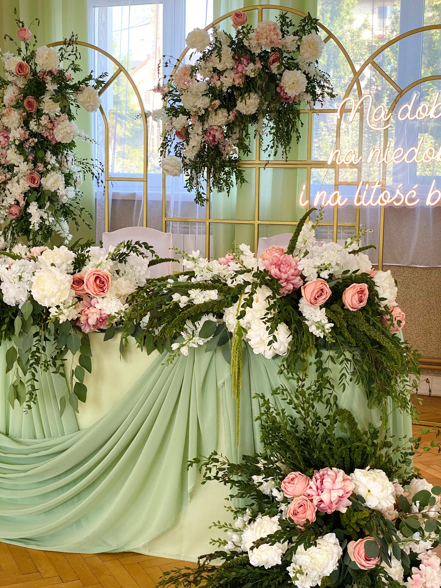 Dekoracja stołu weselnego z kwiatów w odcieniach bieli i różu, z zielonymi akcentami, na tle złotych łuków i neonowego napisu. Elegancka aranżacja.